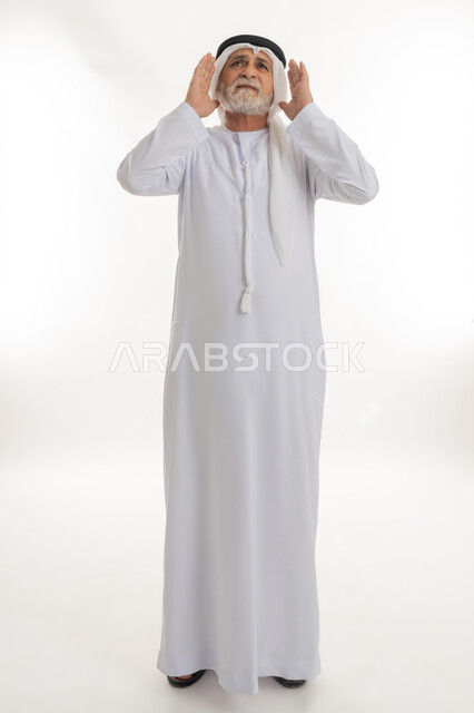 الاستعداد ورفع اليدين والرأس والنظر نحو الأعلى بإيماءات التأمل والخضوع، إشارات وحركات وجه ويدين مختلفة،بورتريه لرجل عربي خليجي إماراتي كبير بالسن يرتدي الكندورة الإماراتية التقليدية والغترة يهم بالصلاة، الجسم بالطول الكامل، خلفية بيضاء