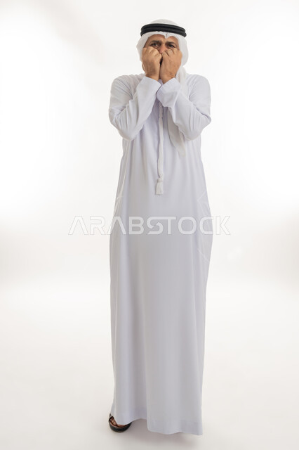 وضع اليدين على الوجه بتعابير الخوف والمفاجأة، بورتريه لرجل عربي خليجي إماراتي كبير بالسن يرتدي الكندورة الإماراتية التقليدية والغترة يغطي وجهه بيديه بإيماءات الذهول، التعبير عن خيبة الأمل والصدمة، الجسم بالطول الكامل، خلفية بيضاء
