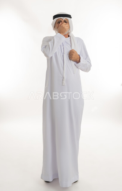النظر إلى الكاميرا والشعور بالاضطراب والتعب، إشارات وحركات وجه ويدين مختلفة تدل على التألم والإحساس بالمرض، بورتريه  لرجل عربي خليجي إماراتي كبير بالسن يرتدي الكندورة الإماراتية التقليدية والغترة يغطي وجهه بيده بإيماءات الحزن، الجسم بالطول الكامل، خلفية بيضاء