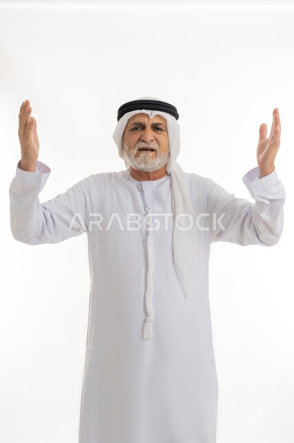 النظر للكاميرا بإيماءات الانزعاج وعدم الرضا، رفع اليدين في الهواء بتعابير الغضب، بورتريه مقرب لرجل عربي خليجي إماراتي كبير بالسن يرتدي الغترة والكندورة الإماراتية يرفع كفيه بتذمر وسخط، خلفية بيضاء