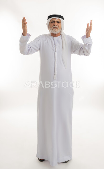 رفع اليدين في الهواء بتعابير الغضب، النظر للكاميرا بإيماءات الانزعاج وعدم الرضا، بورتريه لرجل عربي خليجي إماراتي كبير بالسن يرتدي الغترة والكندورة الإماراتية يرفع كفيه بتذمر وسخط، الجسم بالطول الكامل، خلفية بيضاء