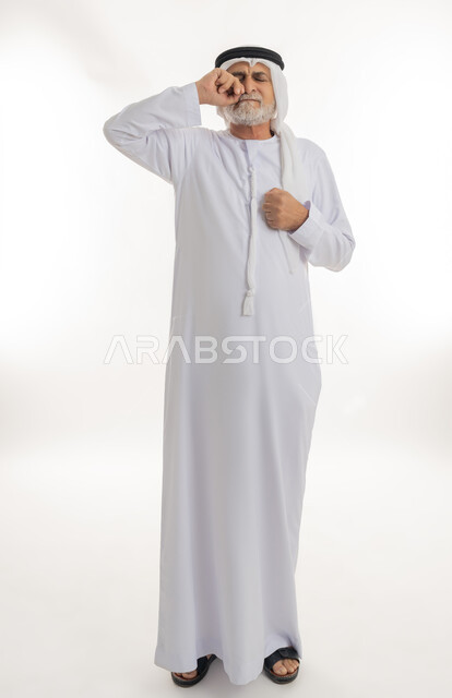 الوقوف والشعور بالاضطراب والتعب، إشارات وحركات وجه ويدين مختلفة تدل على الانزعاج وعدم الرضا، بورتريه لرجل عربي خليجي إماراتي كبير بالسن يرتدي الكندورة الإماراتية التقليدية والغترة يضع قبضته على وجهه بإيماءات التألم، الجسم بالطول الكامل، خلفية بيضاء