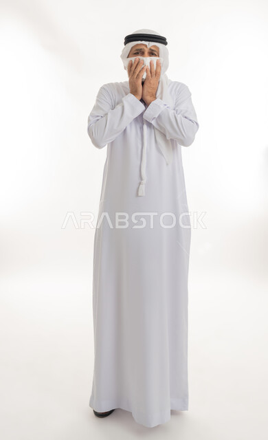 العطاس بشكل متكرر وظهور أعراض الانفلونزا، ضرورة الحصول على الراحة وأخذ العلاج المناسب,   الإصابة بالزكام والتعب والمرض, بورتريه مقرب لرجل كبير السن عربي خليجي اماراتي يرتدي الغترة والكندورة يمسك المنديل بيديه ويغطي أنفه، الجسم بالطول الكامل، خلفية بيضاء