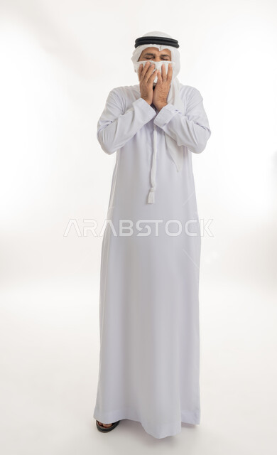 ظهور أعراض الانفلونزا والعطاس بشكل متكرر، ضرورة الحصول على الراحة وأخذ العلاج المناسب,   الإصابة بالزكام والتعب والمرض, بورتريه مقرب لرجل كبير السن عربي خليجي اماراتي يرتدي الغترة والكندورة يمسك المنديل بيديه ويغطي أنفه، الجسم بالطول الكامل، خلفية بيضاء