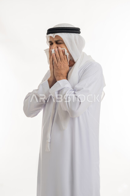 العطاس والإحساس بأعراض الزكام والإصابة بالانفلونزا، إيماءات الألم والتعب والمرض, بورتريه مقرب من الجانب لرجل كبير السن عربي خليجي اماراتي يرتدي الغترة والكندورة يمسك المنديل بيديه ويغطي أنفه، الحاجة للراحة وأخذ العلاج المناسب, خلفية بيضاء
