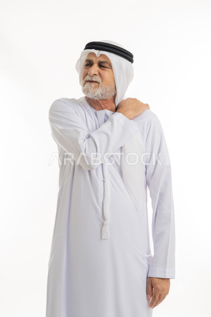 إيماءات الوجه تدل على الاضطراب والتألم من الكتفين، الحاجة للراحة والحصول على العلاج، آلام المفاصل والعظام والعمود الفقري، بورتريه مقرب لرجل كبير السن عربي خليجي إماراتي يرتدي الغترة والكندورة يمسك كتفه بإيماءات الألم, خلفية بيضاء