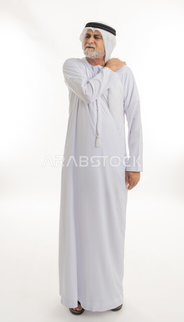الحاجة للراحة والحصول على العلاج، آلام المفاصل والعظام والعمود الفقري، إيماءات الوجه تدل على الاضطراب والتألم من الكتفين، بورتريه لرجل كبير السن عربي خليجي إماراتي يرتدي الغترة والكندورة يمسك كتفه بإيماءات الألم, الجسم بالطول الكامل، خلفية بيضاء