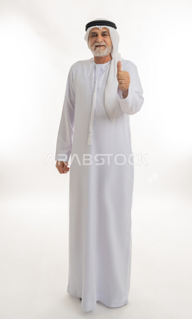 حركات يدين تدل على علامة الجودة والإعجاب، النظر للكاميرا بتعابير السعادة والرضا، بورتريه لرجل عربي خليجي إماراتي كبير بالسن يرتدي الكندورة الإماراتية والغترة يقف ويرفع إبهامه نحو الأعلى  بإيماءات الموافقة، الجسم بالطول الكامل، خلفية بيضاء