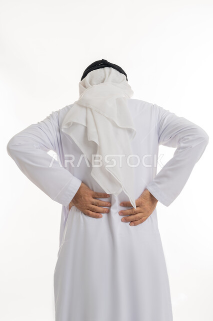 الإحساس بالألم والإصابة بمرض الديسك، آلام الظهر والعمود الفقري والتهاب المفاصل، بورتريه مقرب من الخلف لرجل عربي خليجي إماراتي كبير بالسن يرتدي الكندورة الإماراتية والغترة يقف بصعوبة ويمسك ظهره بيديه بإيماءات المعاناة، خلفية بيضاء
