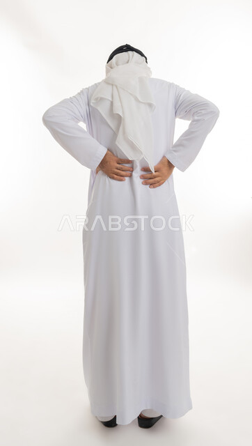 آلام الظهر والعمود الفقري والتهاب المفاصل، بورتريه من الخلف لرجل عربي خليجي إماراتي كبير بالسن يرتدي الكندورة الإماراتية والغترة يقف بصعوبة ويمسك ظهره بيديه بإيماءات المعاناة، الإحساس بالألم والإصابة بمرض الديسك، الجسم بالطول الكامل، خلفية بيضاء