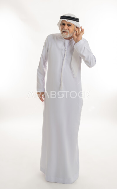 الحاجة لمراجعة طبيب الأذن لتركيب السماعات الطبية, إيماءات التركيز والاستماع لفهم الكلام بشكل واضح، بورتريه لرجل كبير السن عربي خليجي إماراتي يرتدي الغترة والكندورة يمسك أذنه بيده ويعاني من ضعف السمع، الجسم بالطول الكامل، خلفية بيضاء