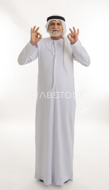 إشارات يدين تدل على علامة الجودة والإعجاب، النظر للكاميرا بتعابير السعادة والرضا، بورتريه  لرجل عربي خليجي إماراتي كبير بالسن يرتدي الكندورة الإماراتية والغترة يقف ويرفع يديه بإيماءات الموافقة، الجسم بالطول الكامل، خلفية بيضاء