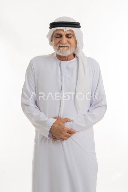 حركات وجه ويدين تدل على الاضطراب والانزعاج، ضرورة الراحة وأخذ العلاج المناسب، الشعور بالمرض ووجع في البطن, بورتريه مقرب لرجل كبير السن عربي خليجي إماراتي يرتدي الغترة والكندورة يضع يديه على بطنه بتعابير التألم, خلفية بيضاء