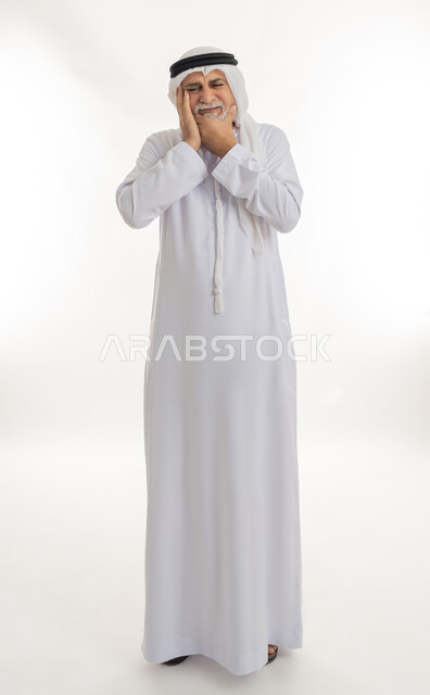 الحاجة للعلاج والذهاب للعيادة السنية، الشعور بالتعب والمرض, بورتريه لرجل كبير السن عربي خليجي إماراتي يرتدي الغترة والكندورة يضع يديه على خده بتعابير الألم, الإحساس بوجع الأسنان والتألم من النخور، الجسم بالطول الكامل، خلفية بيضاء