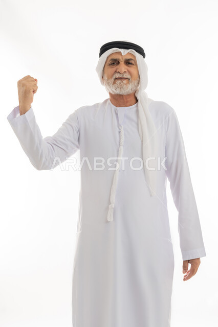 النظر للكاميرا بإيماءات الجدية والوقوف بظهر مستقيم ومشدود، بورتريه مقرب لرجل كبير بالسن عربي خليجي إماراتي يرتدي الكندورة والغترة يرفع يده بتعابير القوة والعزيمة، حركات وإشارات يدين تدل على التحدي والحماس، خلفية بيضاء