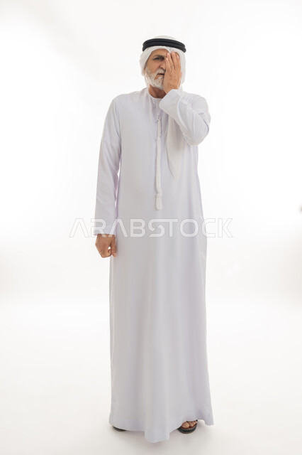 الحاجة إلى الذهاب للطبيب والراحة، إيماءات التألم والشعور بالاضطراب، بورتريه لرجل عربي خليجي إماراتي كبير بالسن يرتدي الكندورة والغترة يضع يده على عينه وينظر إلى الكاميرا بتعابير الألم، الجسم بالطول الكامل، خلفية بيضاء