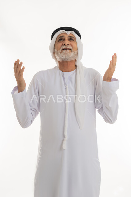 Supplication and raising hands to the sky, close-up portrait of an elderly Emirati Gulf Arab man wearing a kandura and ghutra raising his hands and looking up with expressions of supplication to Allah Almighty, worship and repeating supplications with gestures of submission, white background