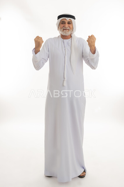 حركات وإشارات تدل على العزيمة والقوة، رفع اليدين بإيماءات الحماس والتشجيع، بورتريه لرجل كبير بالسن عربي خليجي إماراتي يرتدي الكندورة والغترة يقف بظهر مشدود مستقيم وينظر للكاميرا بإيماءات الفرح والسعادة، الجسم بالطول الكامل، خلفية بيضاء