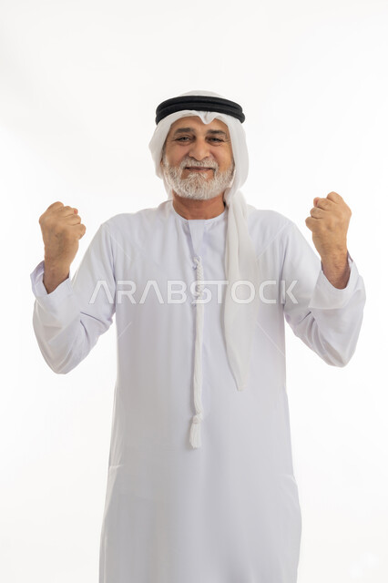رفع اليدين بإيماءات الحماس والتشجيع، بورتريه مقرب لرجل كبير بالسن عربي خليجي إماراتي يرتدي الكندورة والغترة يقف بظهر مشدود مستقيم وينظر للكاميرا بإيماءات الفرح والسعادة، حركات وإشارات تدل على العزيمة والقوة، خلفية بيضاء