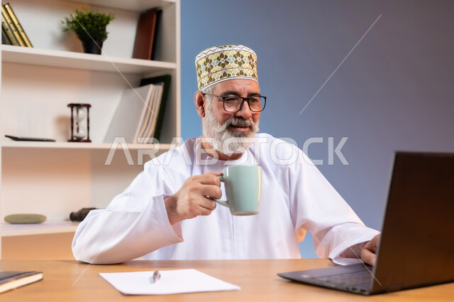 مفهوم ادارة الاعمال عن بعد، مهن ووظائف مكتبيه، بورتريه لرجل كبير السن عربي خليجي عماني يرتدي الدشداشة والكمه يجلس خلف طاولة خشبية يقوم بتناول المشروبات الساخنه و العمل على جهاز الحاسوب المحمول بتعابير السرور، دمج التقنيات الحديثة مع الحياة العملية, خلفية ملونة
