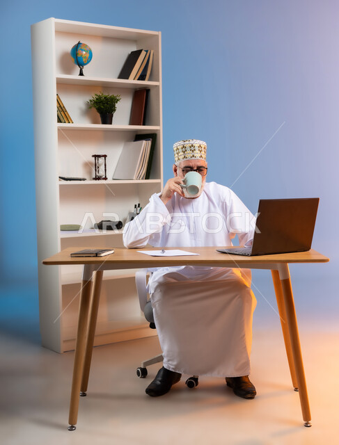 مهن ووظائف مكتبيه، بورتريه لرجل كبير السن عربي خليجي عماني يرتدي الدشداشة والكمه يجلس خلف طاولة خشبية يقوم بتناول المشروبات الساخنه و العمل على جهاز الحاسوب المحمول، دمج التقنيات الحديثة مع الحياة العملية, مفهوم ادارة الاعمال عن بعد، خلفية ملونة