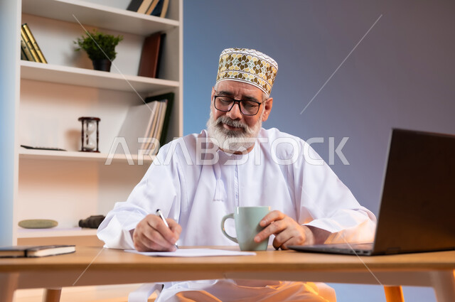 احتساء المشروبات الشتوية الساخنة, مهن ووظائف مكتبيه عمانية, مفهوم ادارة الاعمال عن بعد، بورتريه لرجل كبير السن عربي خليجي عماني يرتدي الدشداشة والكمه العمانية التقليدية يجلس خلف طاولة خشبية يقوم بتدوين الملاحظات