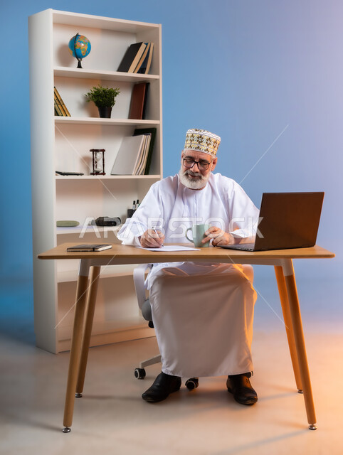 احتساء المشروبات الشتوية الساخنة, مهن ووظائف مكتبيه عمانية, مفهوم ادارة الاعمال عن بعد، بورتريه لرجل كبير السن عربي خليجي عماني يرتدي الدشداشة والكمه العمانية التقليدية يجلس خلف طاولة خشبية يقوم بتدوين الملاحظات
