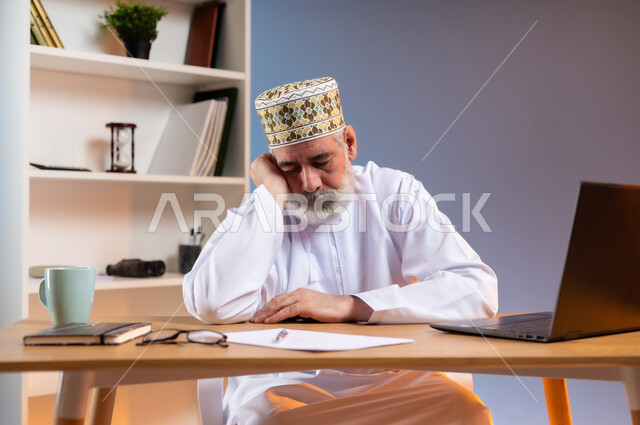 الإحساس بالإرهاق و الحاجه الى النوم والراحه, بورتريه لرجل كبير السن عربي خليجي عماني يرتدي الدشداشة والكمه العمانية التقليدية يجلس خلف طاولة خشبية يضع يده على وجهه بايماءات التعب والنعاس, خلفية ملونة
