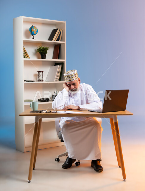 الإحساس بالإرهاق و الحاجه الى النوم والراحه, بورتريه لرجل كبير السن عربي خليجي عماني يرتدي الدشداشة والكمه العمانية التقليدية يجلس خلف طاولة خشبية يضع يده على وجهه بايماءات التعب والنعاس, خلفية ملونة