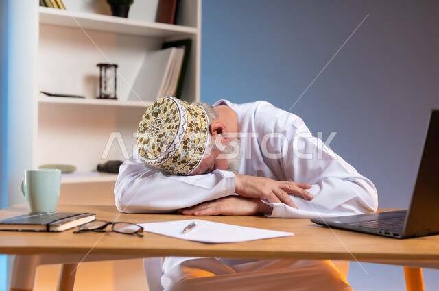 النوم على طاولة العمل، ايماءات النعاس والتعب والإرهاق، استخدام التقنيات الحديثة المتطورة في الحياة العملية، بورتريه مقرب لرجل كبير السن عربي خليجي عماني يرتدي الدشداشة والكمة يقوم بأخذ قسط من الراحة, خلفية ملونة