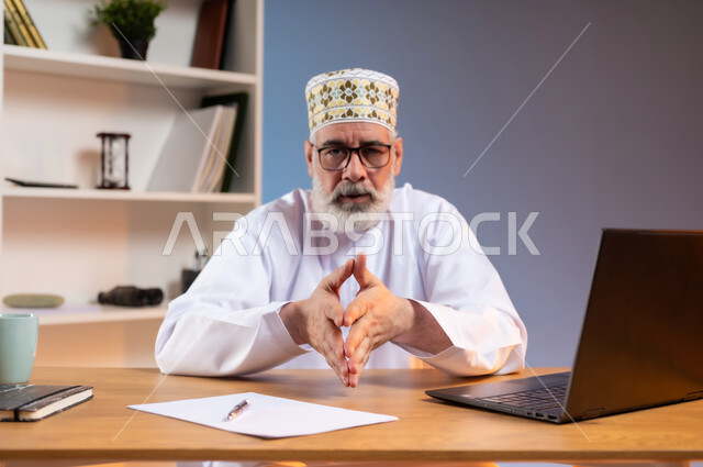 محاولة اخفاء التوتر وتخفيف الضغط النفسي, بورتريه لرجل عربي خليجي عماني يرتدي الدشداشة والكمه العمانية التقليدية يجلس خلف طاولة خشبية يقوم بملامسة أطراف الأصابع بعضها ببعض وينظر إلى الكاميرا بإيماءات الثقة بالنفس، مفهوم الذكاء والإرادة القوية، خلفية ملونة