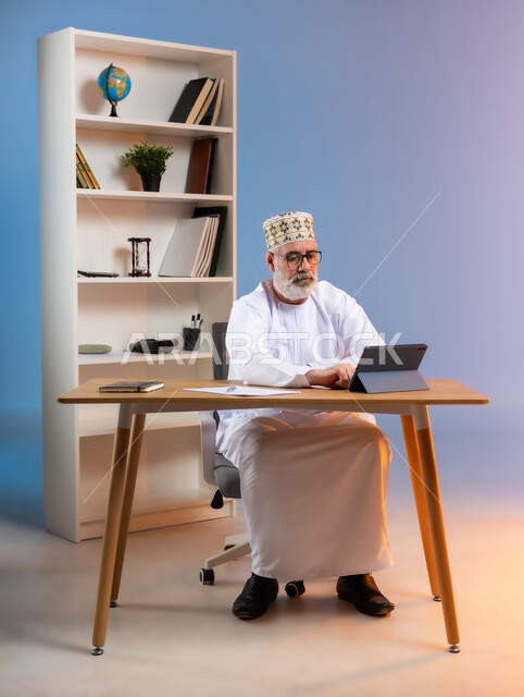 استخدام التقنيات الحديثة في العمل عبر جهاز التابليت، بورتريه لرجل عربي خليجي عماني يرتدي الدشداشة والكمه العمانية التقليدية يجلس خلف طاولة خشبية يقوم باجراء عمله عبر الانترنت, خلفية ملونة