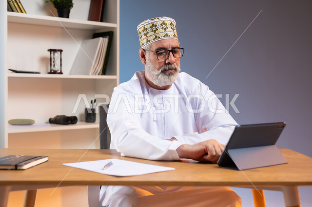 دمج التقنيات الحديثة المتطورة بالحياة العملية، بورتريه لرجل كبير السن عربي خليجي عماني يرتدي الدشداشة والكمه العمانية التقليدية يجلس خلف طاولة خشبية يقوم بالنقر على جهاز التابليت, خلفية ملونة
