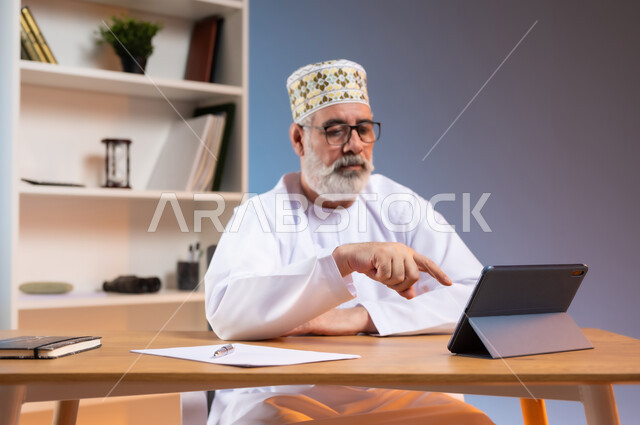 استخدام التقنيات الحديثة والمتطورة في العمل، بورتريه لرجل كبير السن عربي خليجي عماني يرتدي الدشداشة والكمه العمانية التقليدية يجلس خلف طاولة خشبية يشير باصبع السبابة الى جهاز التابليت, خلفية ملونة