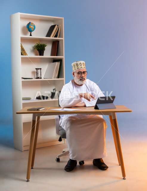 استخدام التقنيات الحديثة والمتطورة في العمل، بورتريه لرجل كبير السن عربي خليجي عماني يرتدي الدشداشة والكمه العمانية التقليدية يجلس خلف طاولة خشبية يشير باصبع السبابة الى جهاز التابليت, خلفية ملونة