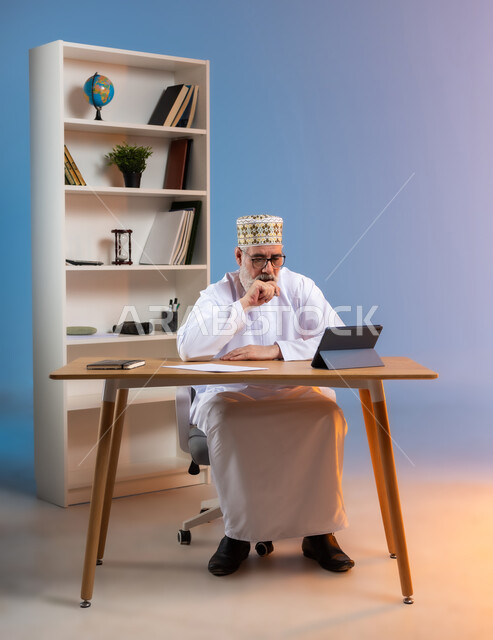 استخدام التقنيات الحديثة والمتطورة في العمل، بورتريه لرجل كبير السن عربي خليجي عماني يرتدي الدشداشة والكمه العمانية التقليدية يجلس خلف طاولة خشبية يضع يده على ذقنه ينظر الى جهاز التابليت بايماءات التعب, خلفية ملونة