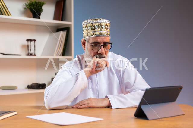 استخدام التقنيات الحديثة والمتطورة في العمل، بورتريه لرجل كبير السن عربي خليجي عماني يرتدي الدشداشة والكمه العمانية التقليدية يجلس خلف طاولة خشبية يضع يده على ذقنه ينظر الى جهاز التابليت بايماءات التعب, خلفية ملونة
