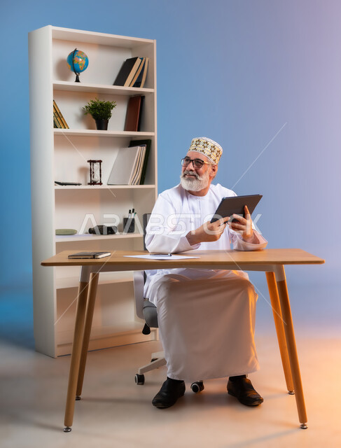 مفهوم ادارة الاعمال عن بعد, استخدام التقنيات الحديثة والمتطورة في العمل، بورتريه لرجل كبير السن عربي خليجي عماني يرتدي الدشداشة والكمه العمانية التقليدية يجلس خلف طاولة خشبية يحمل بيديه جهاز التابليت ينظر الى شيء ما بايماءات السعادة, خلفية ملونة