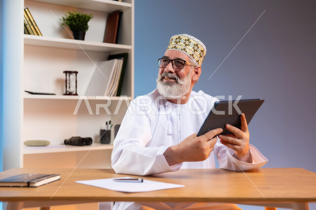 مفهوم ادارة الاعمال عن بعد, استخدام التقنيات الحديثة والمتطورة في العمل، بورتريه مقرب لرجل كبير السن عربي خليجي عماني يرتدي الدشداشة والكمه العمانية التقليدية يجلس خلف طاولة خشبية يحمل بيديه جهاز التابليت ينظر الى شيء ما بايماءات السعادة, خلفية ملونة