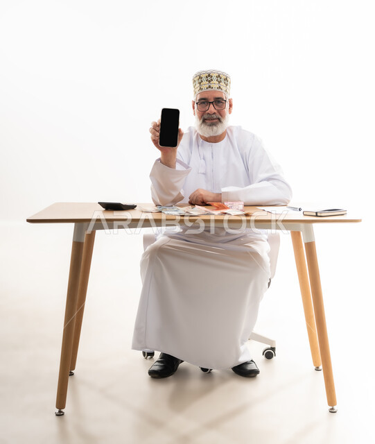 استخدام الأجهزة والتقنيات الحديثة، مهن و أعمال إدارية مكتبية، بورتريه لرجل كبير السن عربي خليجي عماني يرتدي الدشداشة والكمه يجلس خلف طاولة خشبية يمسك بيده الهاتف المحمول يقوم بعرض شاشة سوداء فارغة, خلفية بيضاء