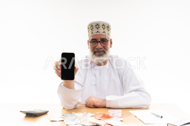 استخدام الأجهزة والتقنيات الحديثة، مهن و أعمال إدارية مكتبية، بورتريه لرجل كبير السن عربي خليجي عماني يرتدي الدشداشة والكمه يجلس خلف طاولة خشبية يمسك بيده الهاتف المحمول يقوم بعرض شاشة سوداء فارغة, خلفية بيضاء