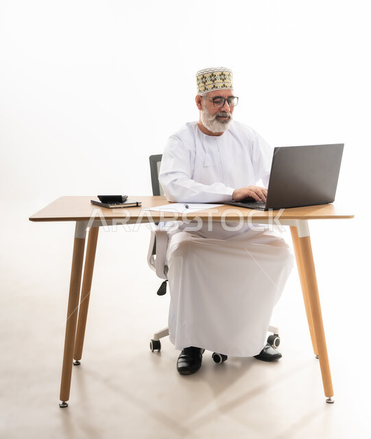 استخدام التقنيات الحديثة والمتطورة في العمل، بورتريه لرجل عربي خليجي عماني يرتدي الدشداشة والكمه العمانية التقليدية يجلس خلف طاولة خشبية يعمل على الحاسوب المحمول, خلفية بيضاء