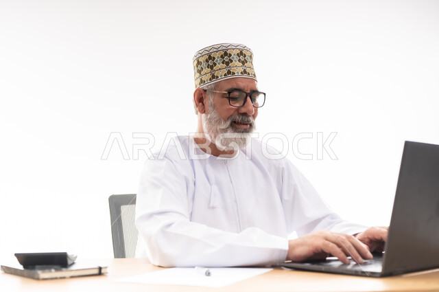 استخدام التقنيات الحديثة والمتطورة في العمل، بورتريه لرجل عربي خليجي عماني يرتدي الدشداشة والكمه العمانية التقليدية يجلس خلف طاولة خشبية يعمل على الحاسوب المحمول, خلفية بيضاء