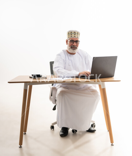 النظر الى الكاميرا بايماءات الثقة بالنفس, استخدام التقنيات الحديثة والمتطورة في العمل، بورتريه لرجل عربي خليجي عماني يرتدي الدشداشة والكمه العمانية التقليدية يجلس خلف طاولة خشبية يعمل على الحاسوب المحمول, خلفية بيضاء