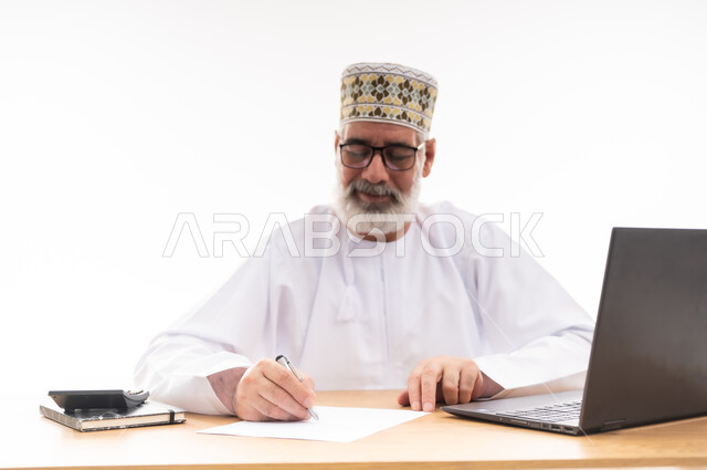 مهن ووظائف مكتبية, استخدام التقنيات الحديثة والمتطورة في العمل، بورتريه لرجل عربي خليجي عماني يرتدي الدشداشة والكمه العمانية التقليدية يجلس خلف طاولة خشبية يقوم بتدوين الملاحظات, خلفية بيضاء