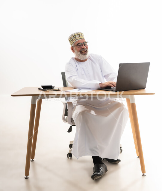 تعابير الضحك والسرور, استخدام التقنيات الحديثة والمتطورة في العمل، بورتريه لرجل عربي خليجي عماني يرتدي الدشداشة والكمه العمانية التقليدية يجلس خلف طاولة خشبية ينظر الى الحاسوب المحمول بايماءات الفرح, خلفية بيضاء