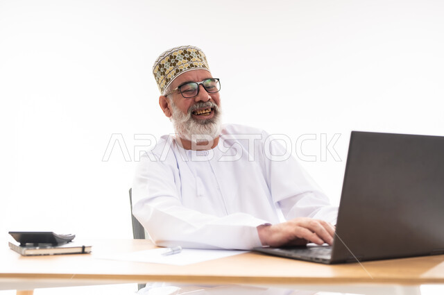 تعابير الضحك والسرور, استخدام التقنيات الحديثة والمتطورة في العمل، بورتريه لرجل عربي خليجي عماني يرتدي الدشداشة والكمه العمانية التقليدية يجلس خلف طاولة خشبية ينظر الى الحاسوب المحمول بايماءات الفرح, خلفية بيضاء