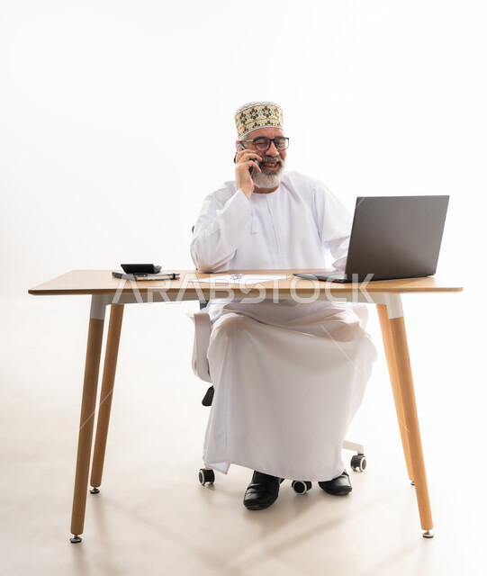 استخدام الأجهزة الحديثة والتقنيات المتطورة، مفهوم ادارة الاعمال عن بعد، مهن ووظائف مكتبيه عمانية, بورتريه لرجل عربي خليجي عماني يرتدي الدشداشة والكمه العمانية التقليدية يقوم باجراء مكالمة هاتفية عبر الهاتف المحمول, خلفية بيضاء