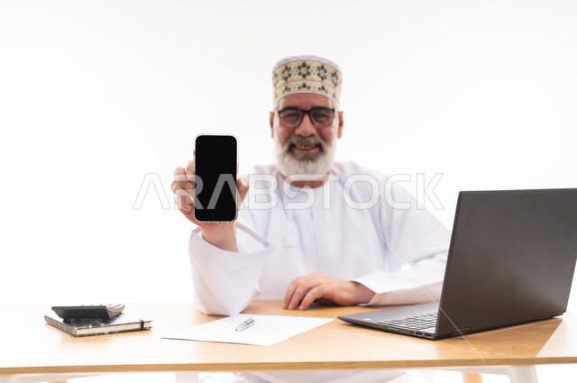 استخدام الأجهزة والتقنيات الحديثة، مهن و أعمال إدارية مكتبية، بورتريه لرجل كبير السن عربي خليجي عماني يرتدي الدشداشة والكمه يجلس خلف طاولة خشبية يمسك بيده الهاتف المحمول يقوم بعرض شاشة سوداء فارغة, خلفية بيضاء
