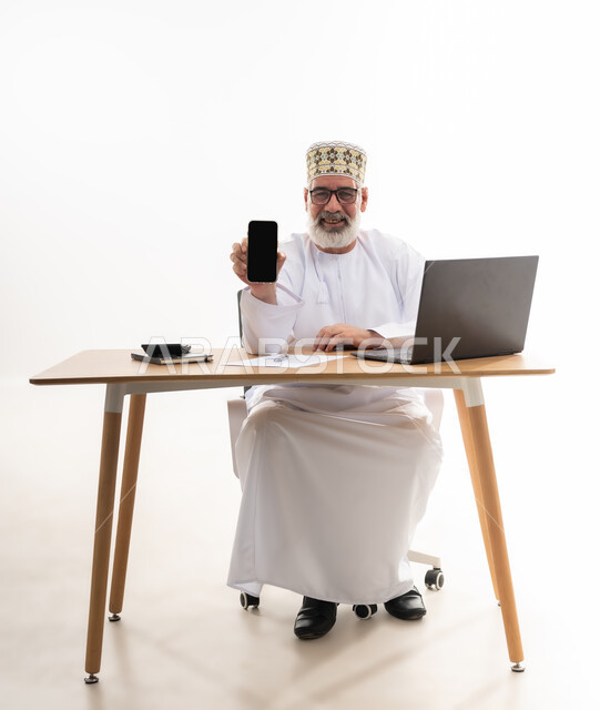 استخدام الأجهزة والتقنيات الحديثة، مهن و أعمال إدارية مكتبية، بورتريه لرجل كبير السن عربي خليجي عماني يرتدي الدشداشة والكمه يجلس خلف طاولة خشبية يمسك بيده الهاتف المحمول يقوم بعرض شاشة سوداء فارغة, خلفية بيضاء